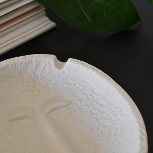 Cenicero de cerámica blanco con relieve de un rostro.