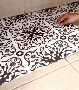 En la imagen se ve un piso en el que se están instalando baldosas de vinilo adhesivas de color blanco con dibujos azul marino. En la esquina derecha una mano pega una baldosa al piso.